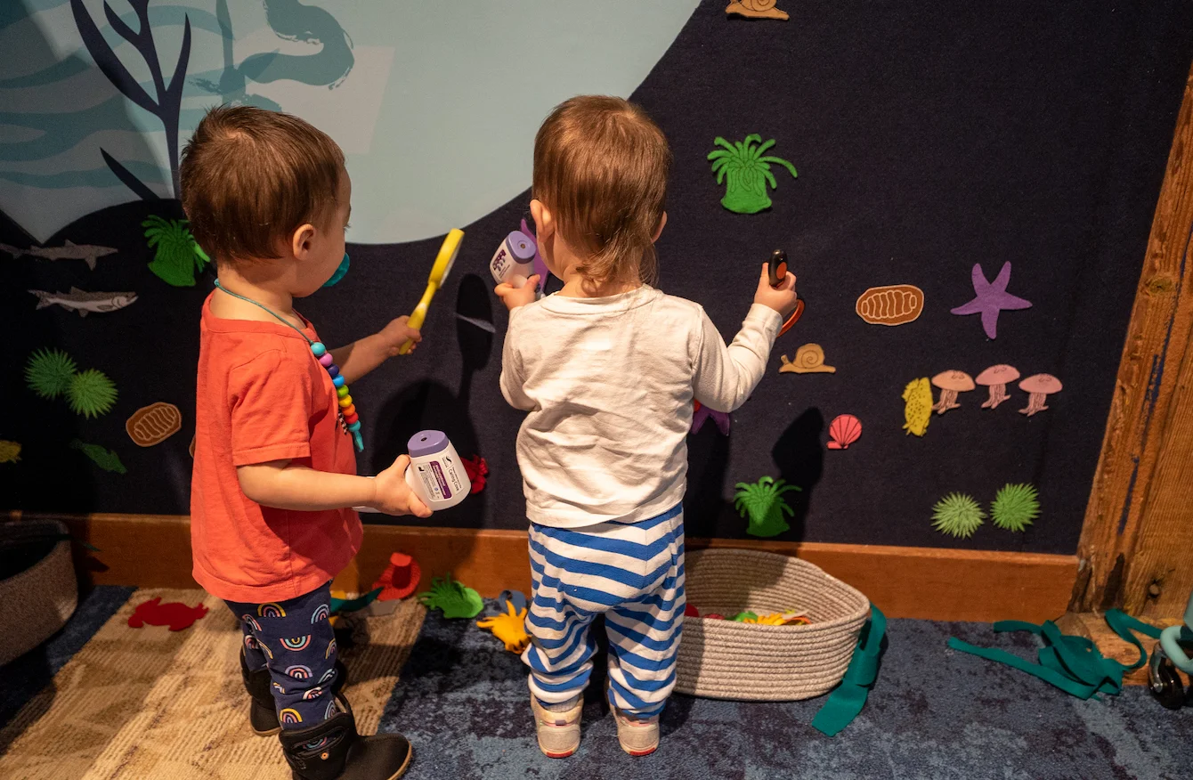 Two young toddlers stand at an activity wall, which is part of the Caring Cove space at the Seattle Aquarium, placing felt animals and objects on the wall.
